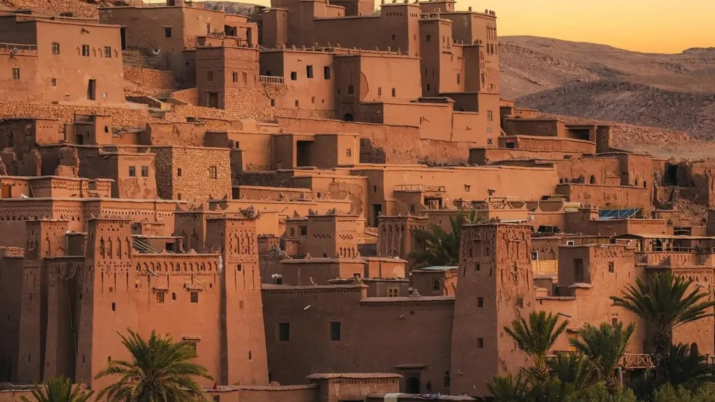 A cluster of traditional earthen buildings stacked on a hillside, surrounded by palm trees in a desert setting. Ait Benhaddou, a highlight on a Morocco itinerary, one week from Fes to Marrakech