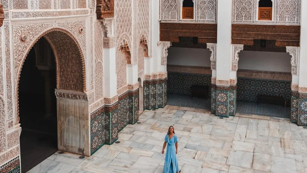 A tourist in a light blue dress stands in the marble courtyard of Ben Youssef Madrasa in Marrakech, surrounded by intricate Islamic geometric patterns and carved wooden archesvisited during a one-week Morocco itinerary with a local guide.