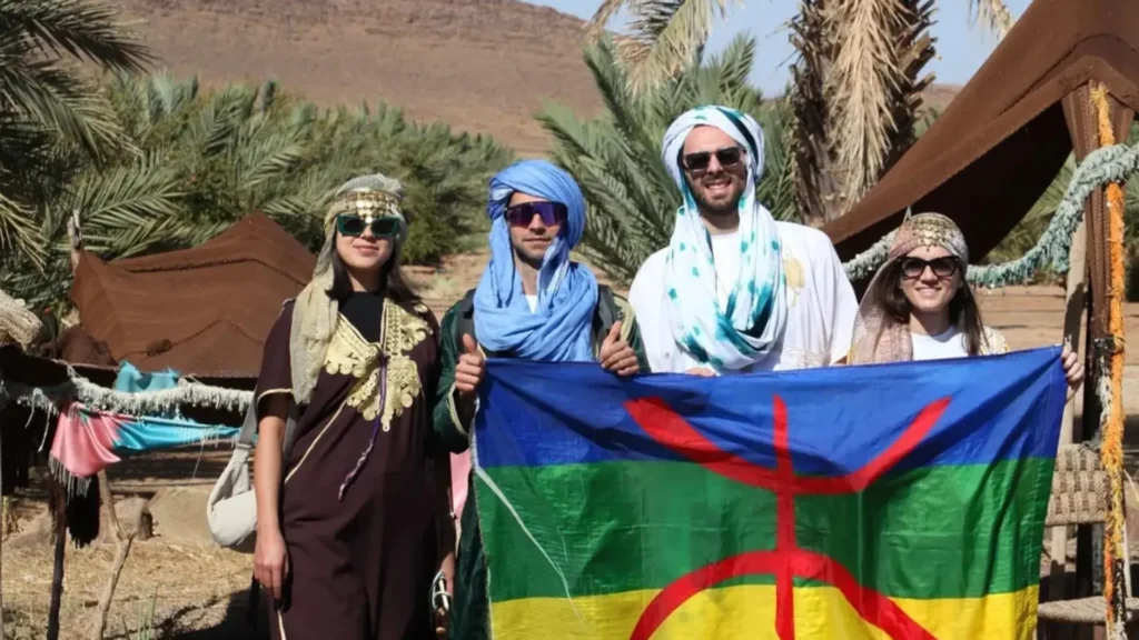 Four tourists wearing traditional Amazigh clothing and holding the Amazigh flag in a Moroccan desert setting with tents and palm trees in the background. This is one of the stops on a Morocco itinerary one week.