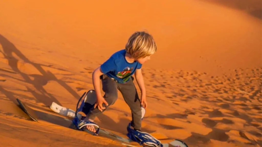 A child sandboarding in Merzouga Desert wearing a blue shirt among golden sand dunes during a one-week Morocco tour