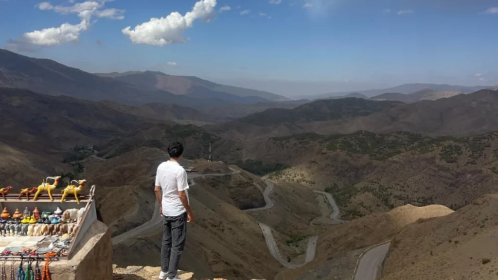 A panoramic view of the winding mountain roads in the High Atlas, with a traveler admiring the scenery from the Tizi n’Tichka Pass during a 3 days desert trip from Marrakech to Fes.