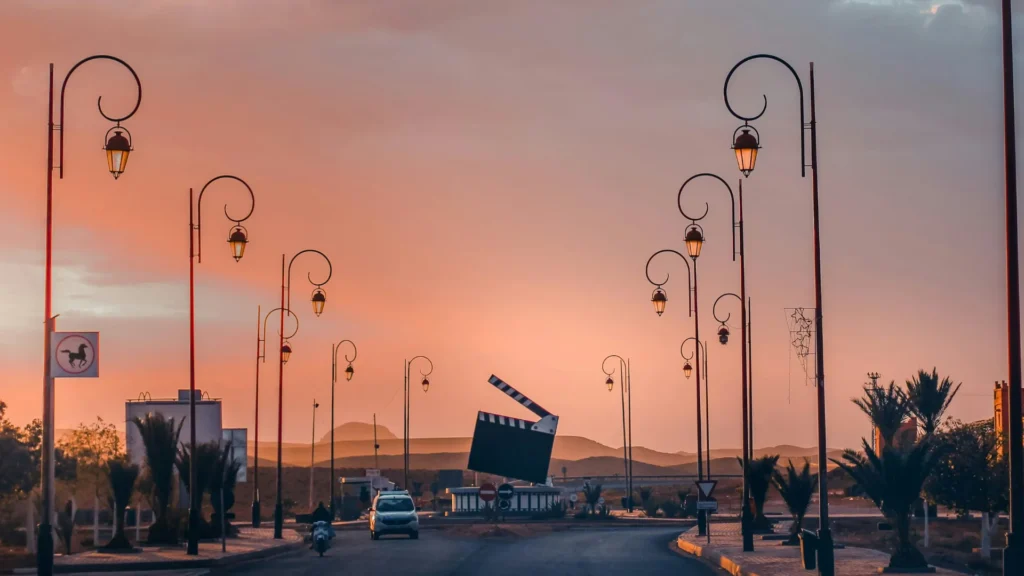 A large film clapperboard sculpture at the center of a roundabout in Ouarzazate, surrounded by palm trees and ornate street lamps, with mountains in the background under a pastel sky.