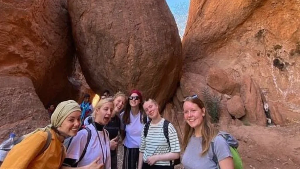A group of tourists posing in front of dramatic reddish-brown rock formations in Dades Valley under a clear blue sky during a one-week itinerary across Morocco from Fes to Marrakech through the desert
