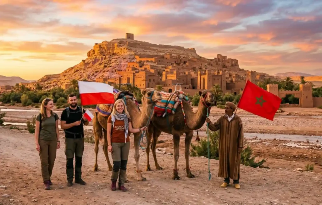 A group of Polish travelers with camels and their local guide holding the flags of Poland and Morocco at the UNESCO World Heritage site of Aït Benhaddou, illustrating the best way to enjoy Morocco Desert Trips and Marrakech Desert Tours.