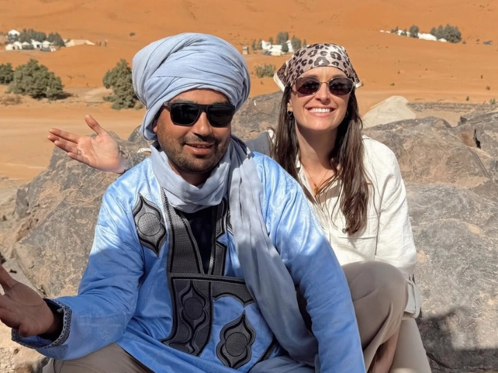 A local man from the Berber Morocco people in a light blue traditional robe and turban sitting on a rock with a smiling tourist woman in the Merzouga desert.