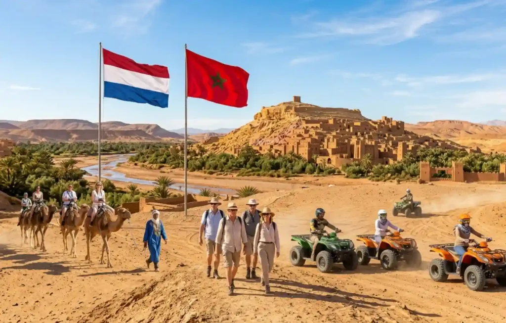 A group of tourists from the Netherlands to Morocco enjoying an outdoor adventure at Ait Ben Haddou, featuring the Dutch and Moroccan flags waving in the foreground. Travelers are shown riding camels in a traditional caravan and driving ATVs across the golden desert sands, with the historic mud-brick ksar and palm-filled riverbed in the background during a sunny day.
