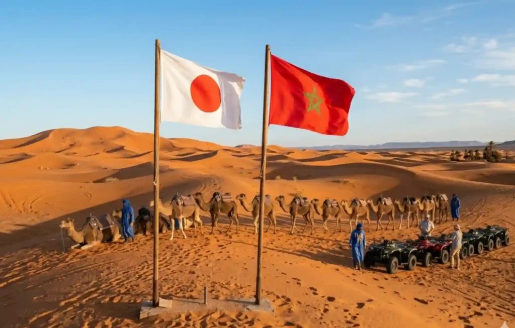 The national flags of Japan and Morocco wave on poles in the Sahara Desert dunes of Merzouga. Below the flags, a caravan of camels and a line of quad bikes are prepared for a desert excursion at sunset, symbolizing a welcoming bridge for those who travel to Morocco from Japan to experience the golden dunes.