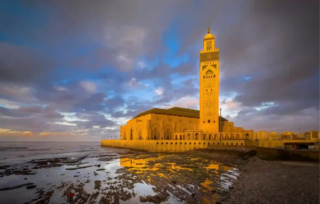 The Hassan II Mosque in Casablanca, Morocco, standing on the edge of the Atlantic Ocean during a golden hour sunset with dramatic clouds. This iconic landmark is a must-see for travelers who travel to Morocco from Japan, featuring the world's tallest minaret reflecting in the coastal tide pools.