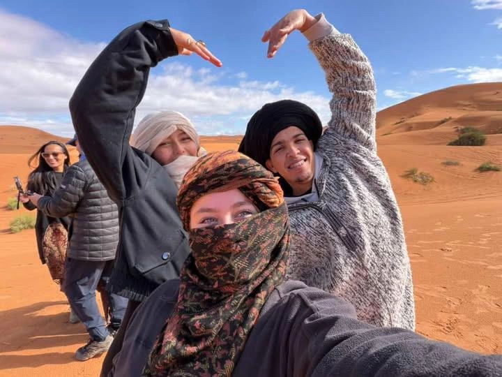 A group of happy travelers and a local guide from the Moroccan people making heart shapes with their hands in the Sahara dunes.