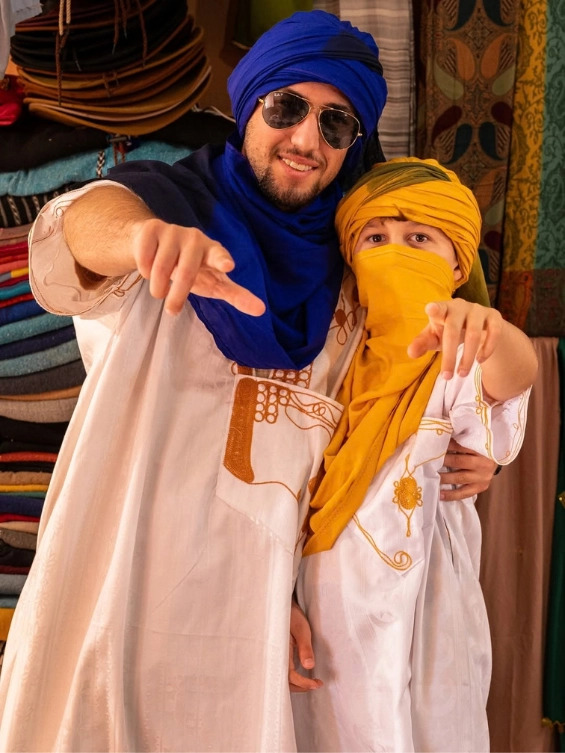 Two Moroccan people, an adult and a child, wearing traditional white djellabas and colorful blue and yellow turbans in the Merzouga desert.