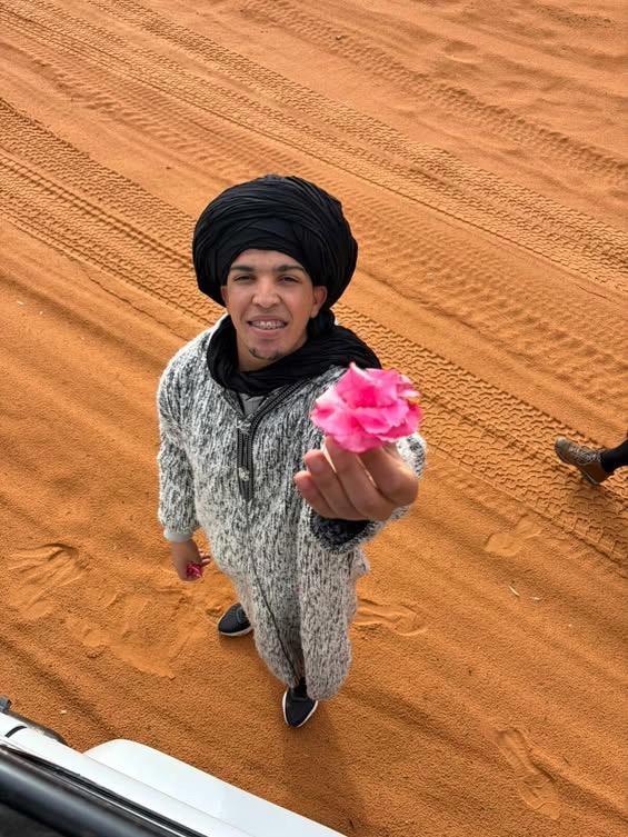 A smiling young man from the Moroccan people wearing a traditional djellaba and black turban, offering a pink flower in the Merzouga desert.