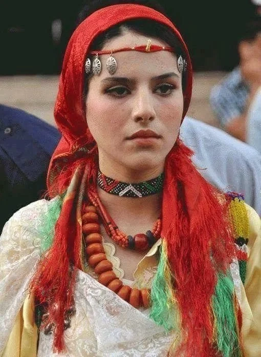 A young woman from the Moroccan people wearing traditional Berber jewelry, a red headscarf, and a vibrant beaded necklace.