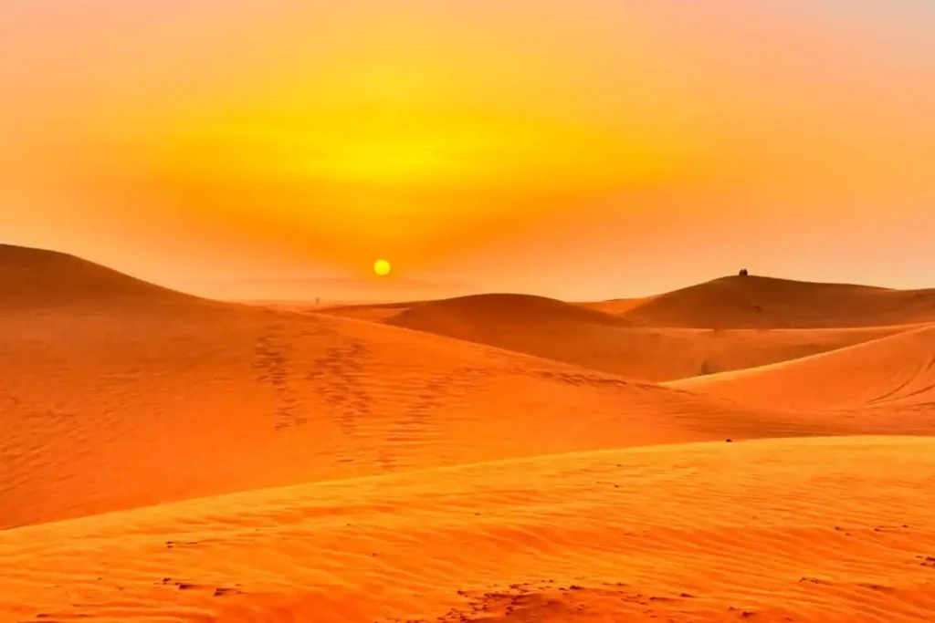 A breathtaking sunset over the vast orange sand dunes of Merzouga with the sun appearing as a small yellow orb on the horizon under a glowing sky illustrating the goal of the long 10 hour drive from Marrakech to Merzouga.