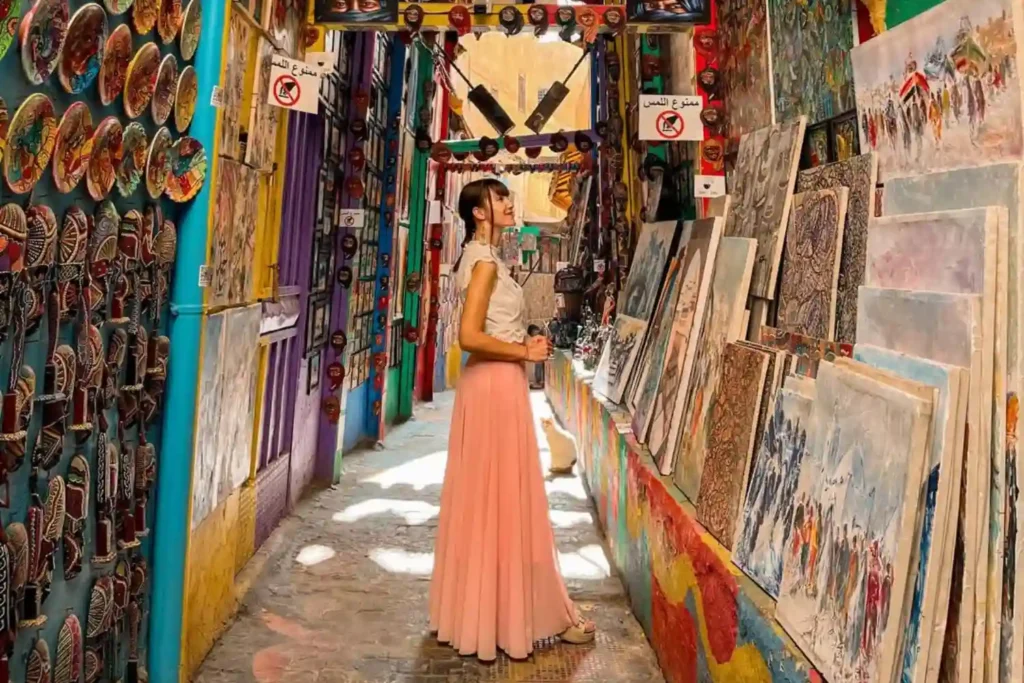 A female traveler in a long pink skirt exploring a colorful outdoor art gallery and souvenir shop in a narrow alley of the Fes Medina, illustrating why is Fes safe for tourists seeking authentic cultural experiences.