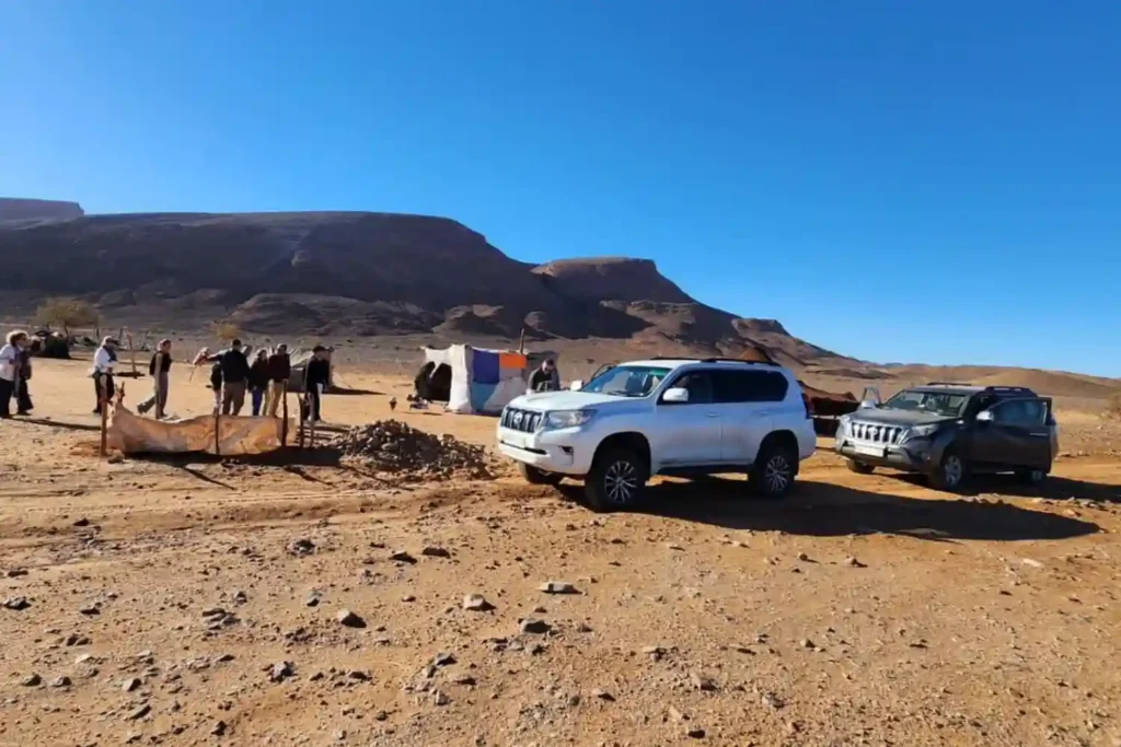 A diverse tour group gathered at a remote Berber desert camp near dramatic mountains, with white and gray Toyota SUVs parked. A local guide shows off ancient fossils, highlighting cultural and historical things to do in the Sahara Desert in Morocco.