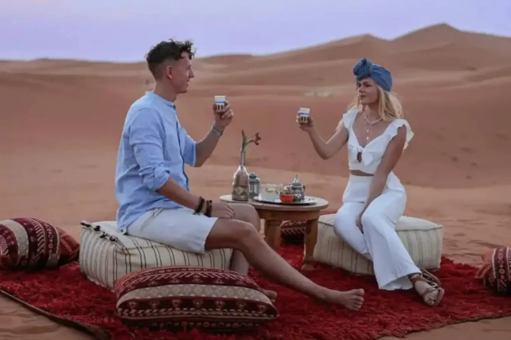 A vibrant travel collage illustrating the ultimate reward for travelers asking how far is Merzouga from Marrakech featuring a group of five friends celebrating with drinks and white table service on a red carpet in the golden dunes of Erg Chebbi at sunset with camels in the background and traditional Moroccan headscarf wrapping.