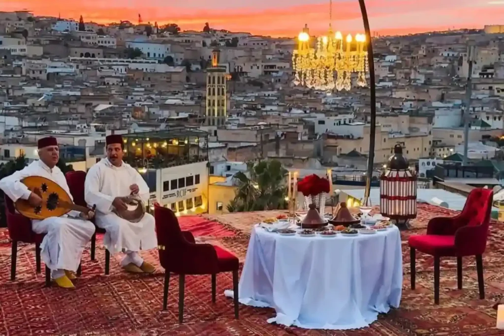 A peaceful rooftop terrace in Fes at sunset featuring a set dinner table with traditional Moroccan tagines and two local musicians playing instruments, overlooking the sprawling ancient Medina.