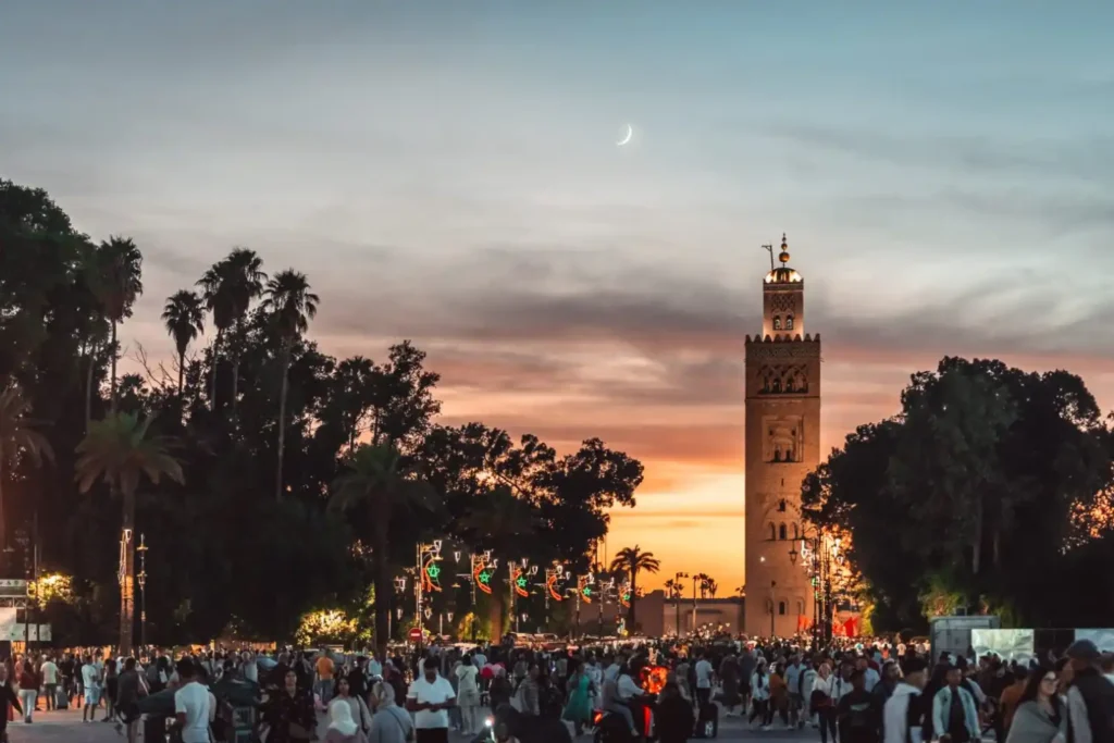 A vibrant evening scene in Marrakech showing a large, diverse crowd of people walking near the illuminated Koutoubia Mosque at sunset, illustrating the busy and well-lit public atmosphere.