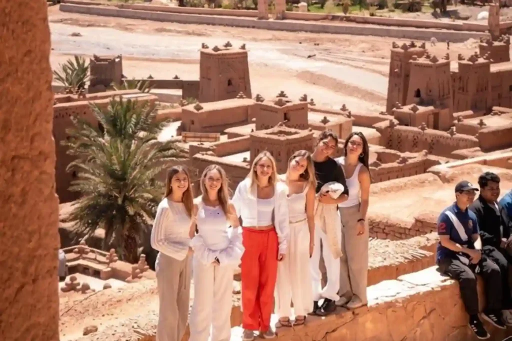 A group of young women travelers posing together at the ancient Ait Benhaddou kasbah in Morocco, wearing modest summer clothing and demonstrating that solo female travel in Morocco is safe, accessible, and highly social.