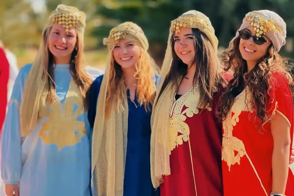 Four smiling female tourists participating in a cultural experience by wearing traditional Moroccan Berber robes (Kaftans) in vibrant colors with ornate gold headpieces, representing respectful solo female travel in Morocco.