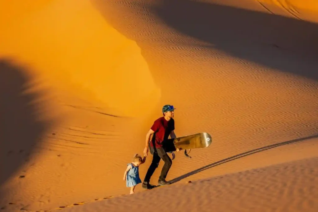 A detailed view of a man and a small child holding hands and ascending a monumental, rippled golden sand dune. The man, wearing a red shirt, blue hat, and dark trousers, carries a snowboard/sandboard, while the child is in a blue dress. The entire scene is bathed in deep golden afternoon light, highlighting the vastness and textures of the dunes during a family tour in the expansive Sahara Desert in Morocco.