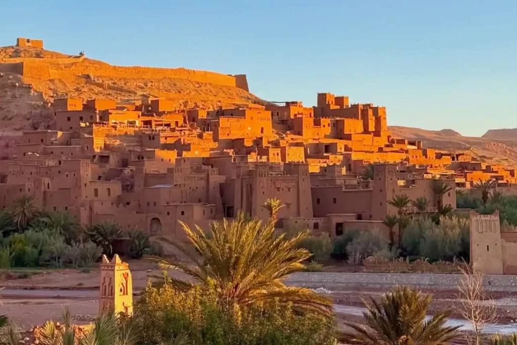 A wide-angle, high-resolution photograph of the ancient earthen clay architecture of the Ksar of Ait Ben Haddou in Morocco, captured during the golden hour sunset. This image showcases the intricate defensive walls, high towers, and palm-fringed riverbed, serving as a primary cultural attraction for travelers booking a Marrakech itinerary 3 Days to explore the desert and Atlas Mountains.