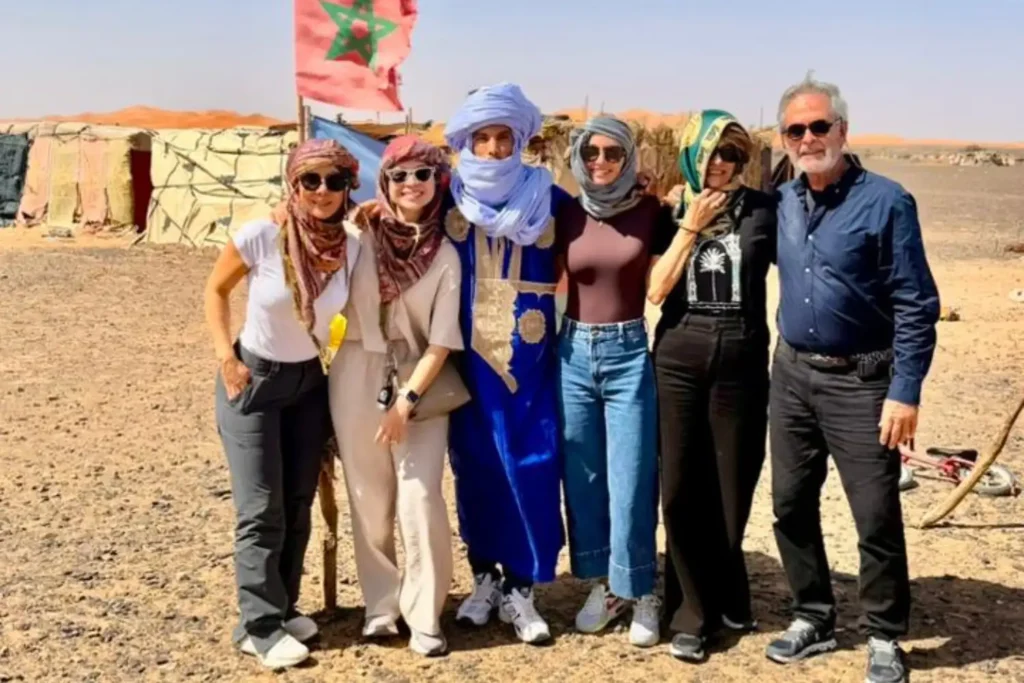 A group of international travelers posing with a local Berber guide in traditional blue robes and a litham turban in front of a nomadic tent camp with the Moroccan flag, a key cultural highlight of a professional 3 day Marrakech itinerary through the Merzouga desert.