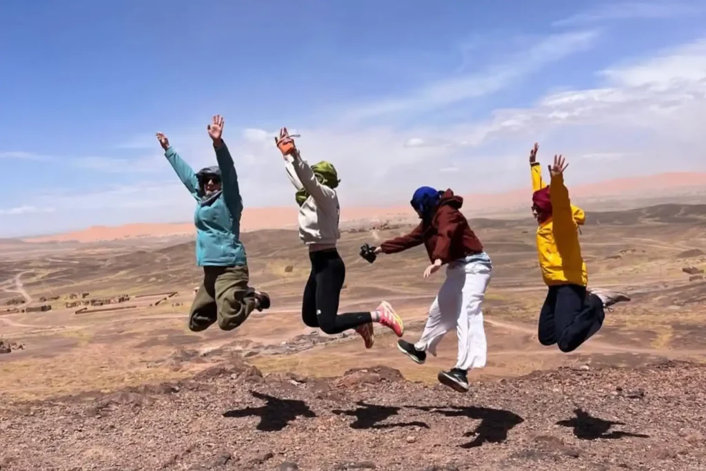 A group of four happy travelers jumping in the air on a rocky mountain overlook with a vast desert valley and orange sand dunes in the background, showcasing the excitement of a professional Marrakech 3 day itinerary.