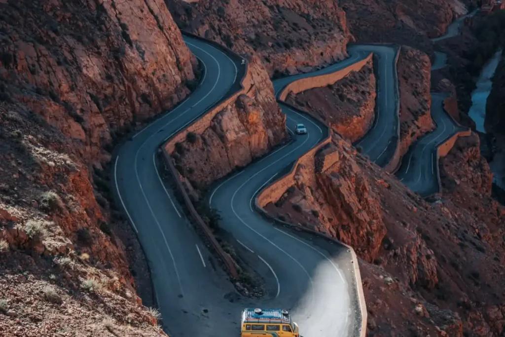 An aerial view of the winding mountain roads and switchbacks of the Tizi n'Tichka pass in the High Atlas Mountains, featuring a yellow transport van navigating the scenic curves at sunset during a professional 3 day Marrakech itinerary.