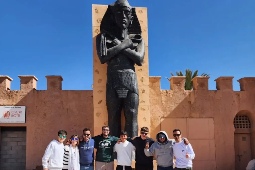 A group of eight travelers posing happily in front of a giant Egyptian pharaoh statue at the Atlas Film Studios in Ouarzazate, a major cinematic landmark often visited during a comprehensive Marrakech itinerary 3 Days through Southern Morocco.