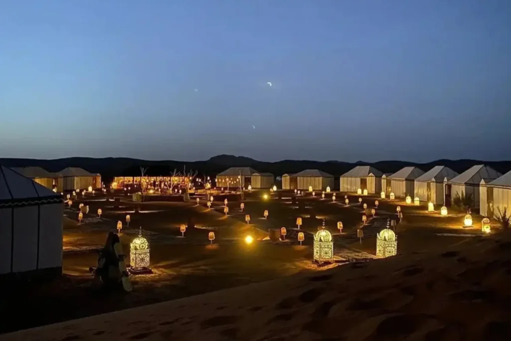 A wide view of a luxury desert camp in Merzouga at twilight, featuring white canvas tents illuminated by dozens of glowing lanterns along the pathways under a deep blue night sky, a premium accommodation option on a Marrakech 3 day itinerary.