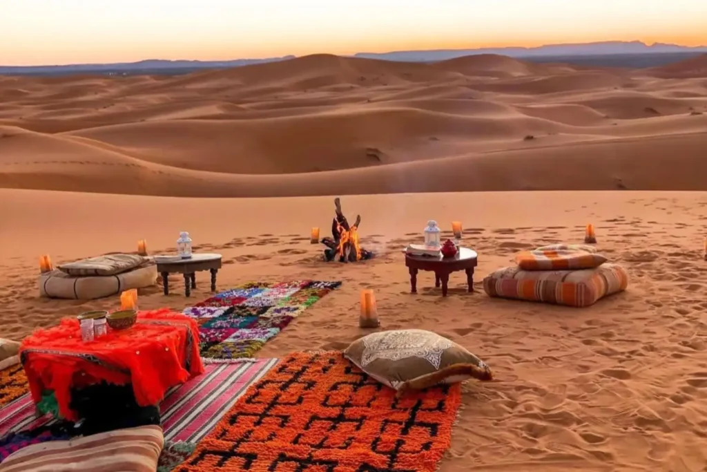 A cozy outdoor desert lounge setup in the Erg Chebbi dunes at sunset, featuring traditional colorful Moroccan rugs, floor cushions, low wooden tables, and a crackling central campfire with views of the rolling sand dunes, a core highlight of a Marrakech itinerary 3 Days.