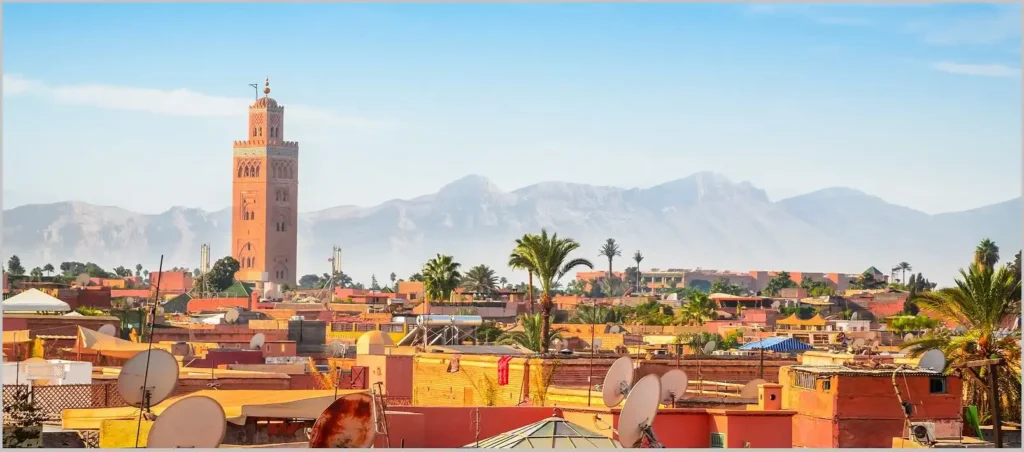 A breathtaking wide-angle panoramic view of the Moroccan skyline answering the question Where is Marrakech by showing the historic Koutoubia Mosque minaret towering over the red sandstone buildings of the ancient Medina, with the majestic, hazy peaks of the High Atlas Mountains serving as a dramatic geographic backdrop under a clear blue sky.
