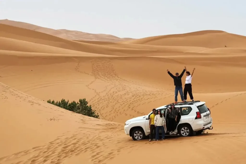A white 4x4 Toyota Land Cruiser parked on a steep orange sand dune in Merzouga, with travelers standing on top of the vehicle and posing next to it, a signature experience of a Marrakech itinerary 3 Days.